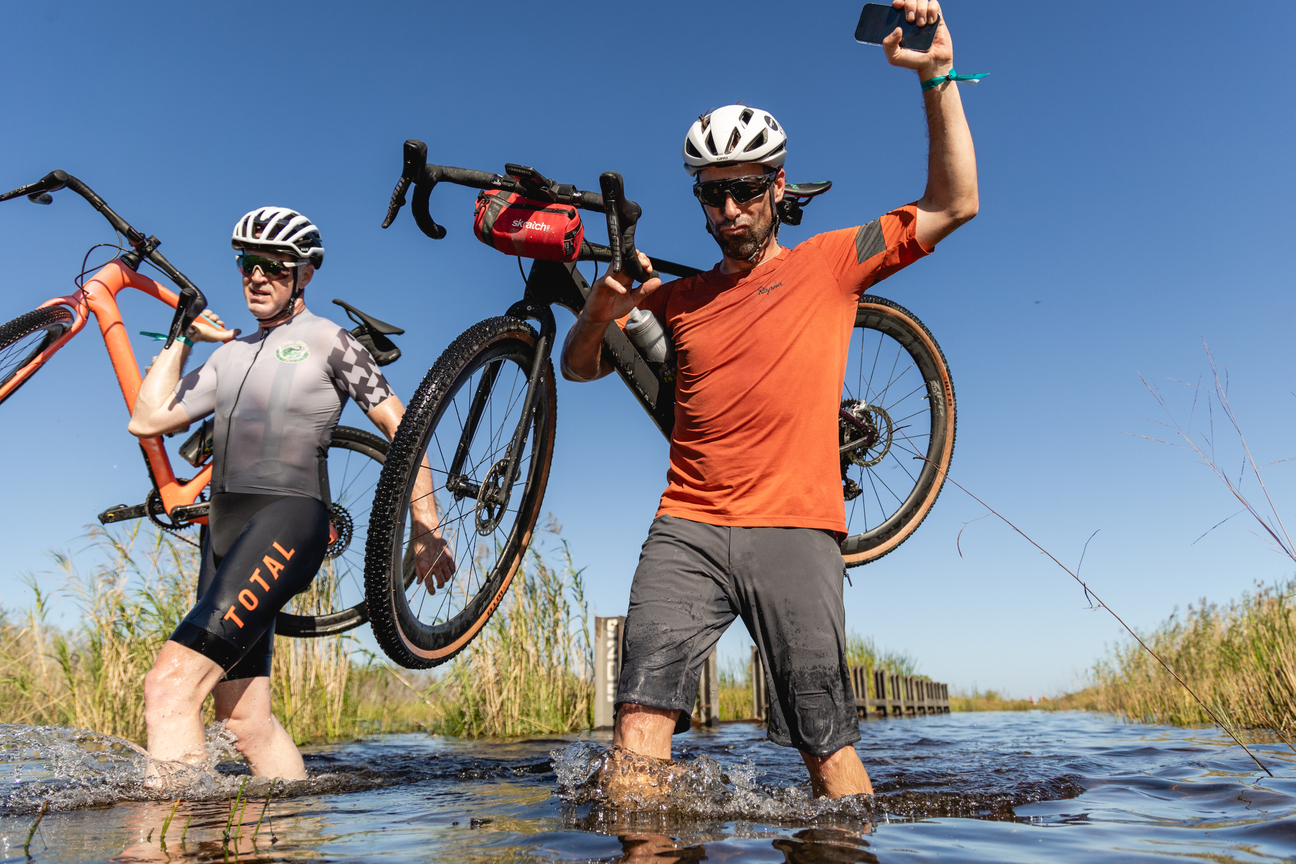 GREG DEWEESE AND TIM JOHNSON CARRYING THEIR BIKES THROUGH A WATER CROSSING AT MACK CYCLE GRAVEL RIDE